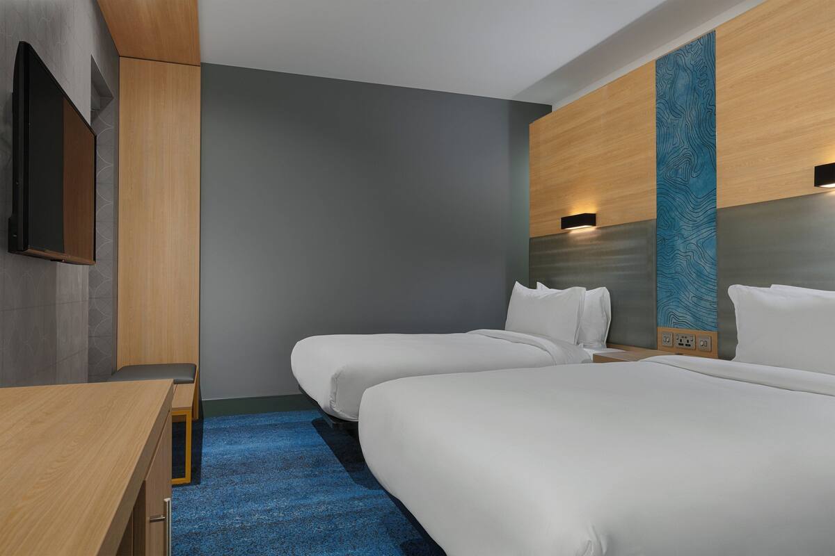 Ein Hotelzimmer mit zwei Einzelbetten, einem Schreibtisch, einem Flachbildfernseher und einem an der Wand befestigten Fernseher.