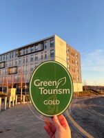 Uma mão segura um sinal circular com o texto 'Green Tourism GOLD', em frente a um edifício moderno com várias janelas.