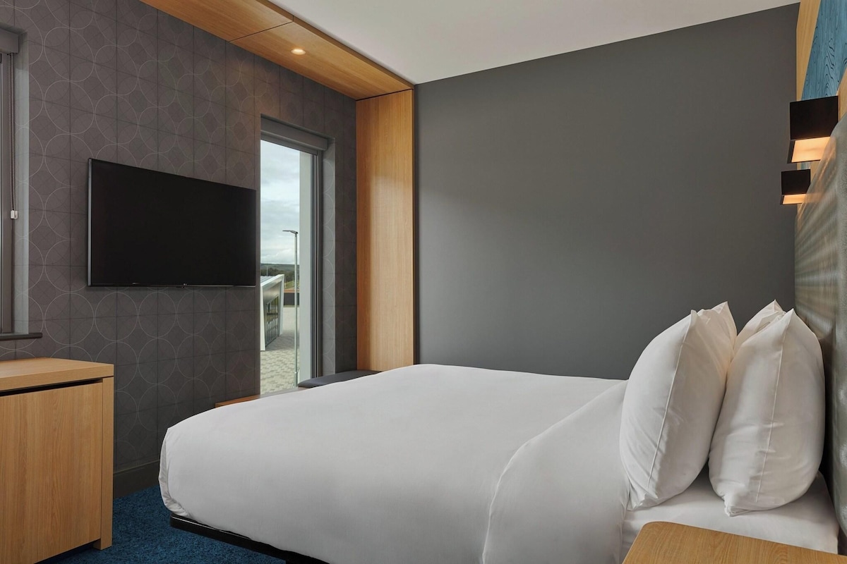Een hotelkamer met een groot bed, een flatscreen televisie aan de muur, een houten nachtkastje en uitzicht op het strand door een glazen deur.