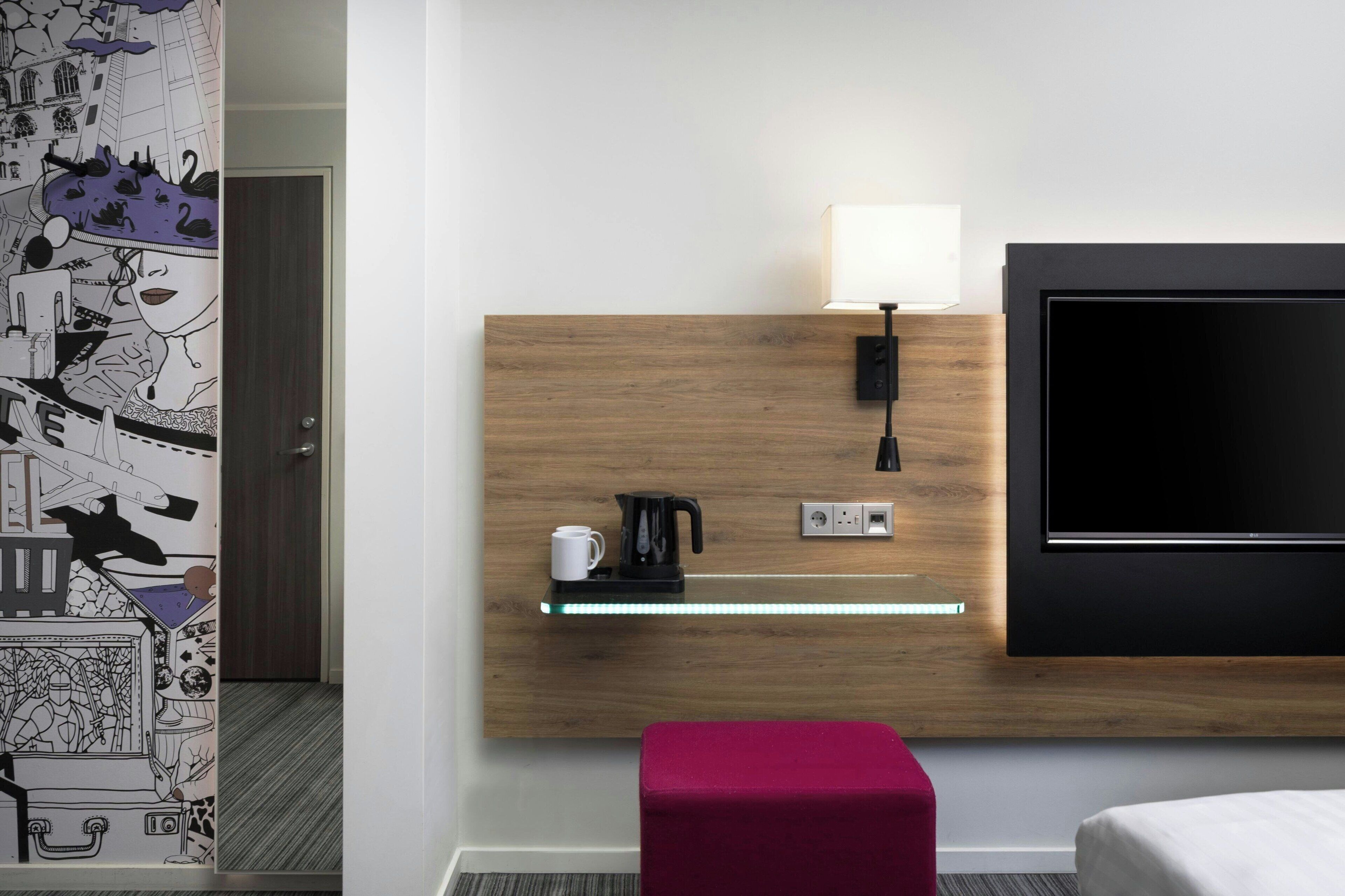 Habitación de hotel con cabecera de madera, televisor de pantalla plana, cafetera y una pequeña repisa con una taza.