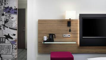 Habitación de hotel con cabecera de madera, televisor de pantalla plana, cafetera y una pequeña repisa con una taza.