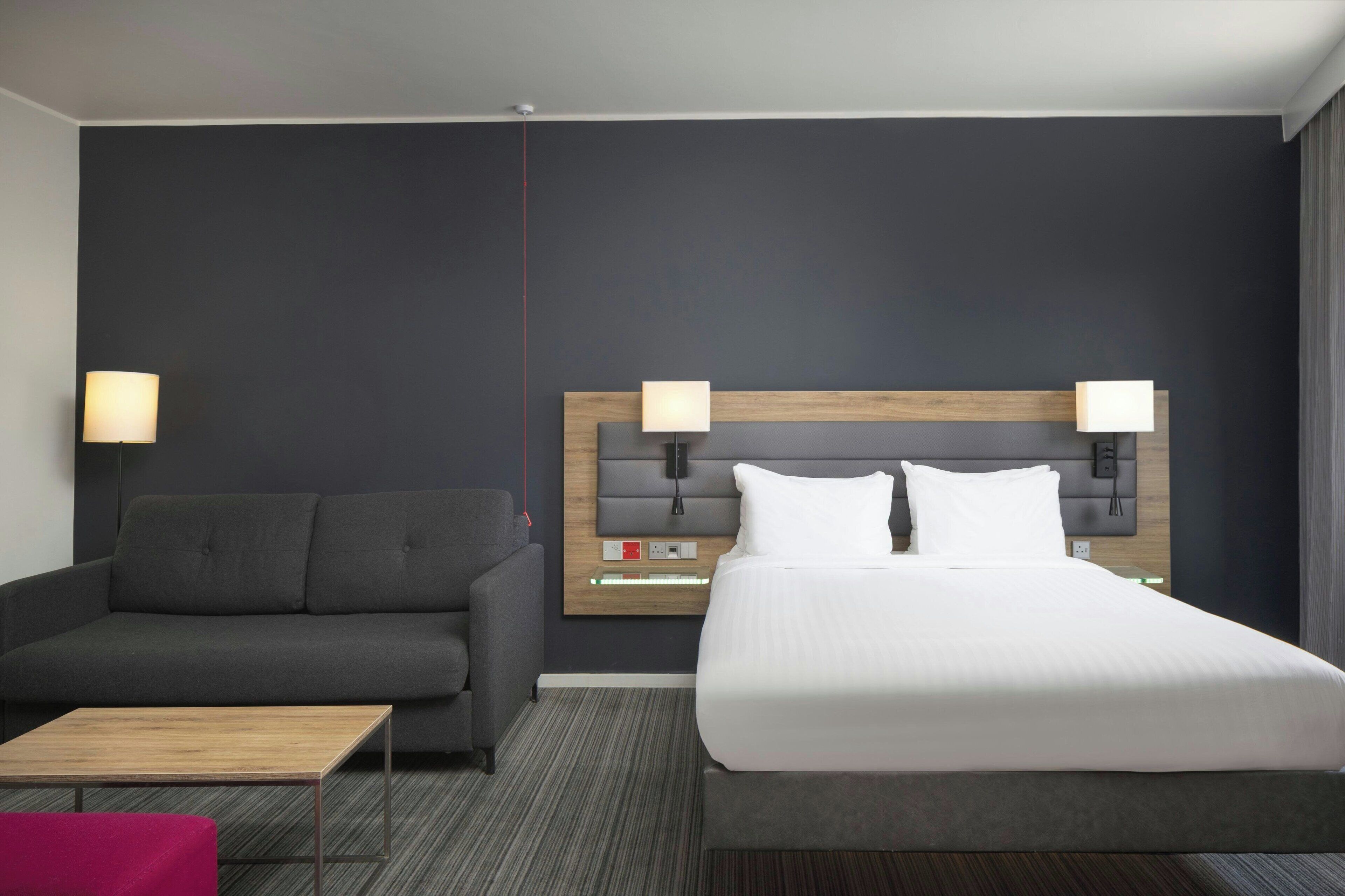 A hotel room with a bed, a sofa, a small table, and two wall-mounted lamps.