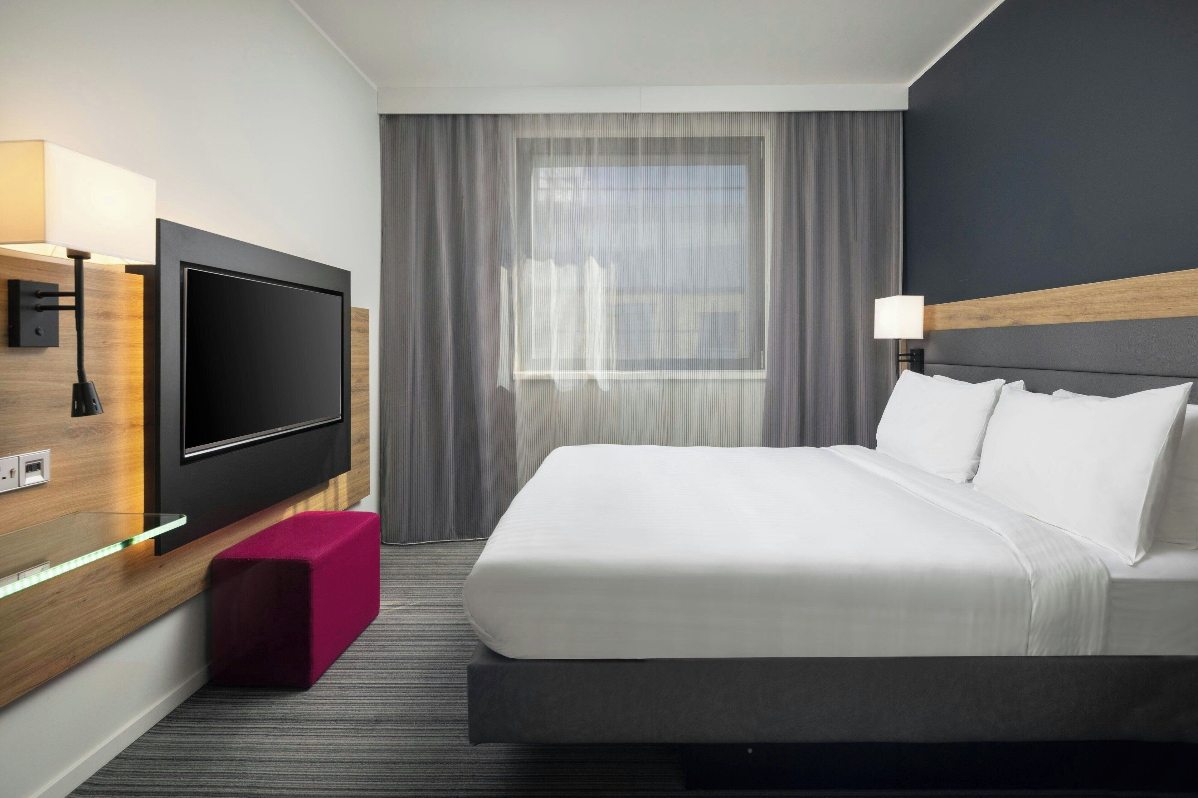 A modern hotel room with a large flat-screen TV mounted on the wall, a bed with white linens, a wooden headboard, a small pink ottoman, and a window with curtains.