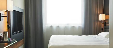 A hotel room with a bed, a TV, a desk with a lamp, and a window with curtains.