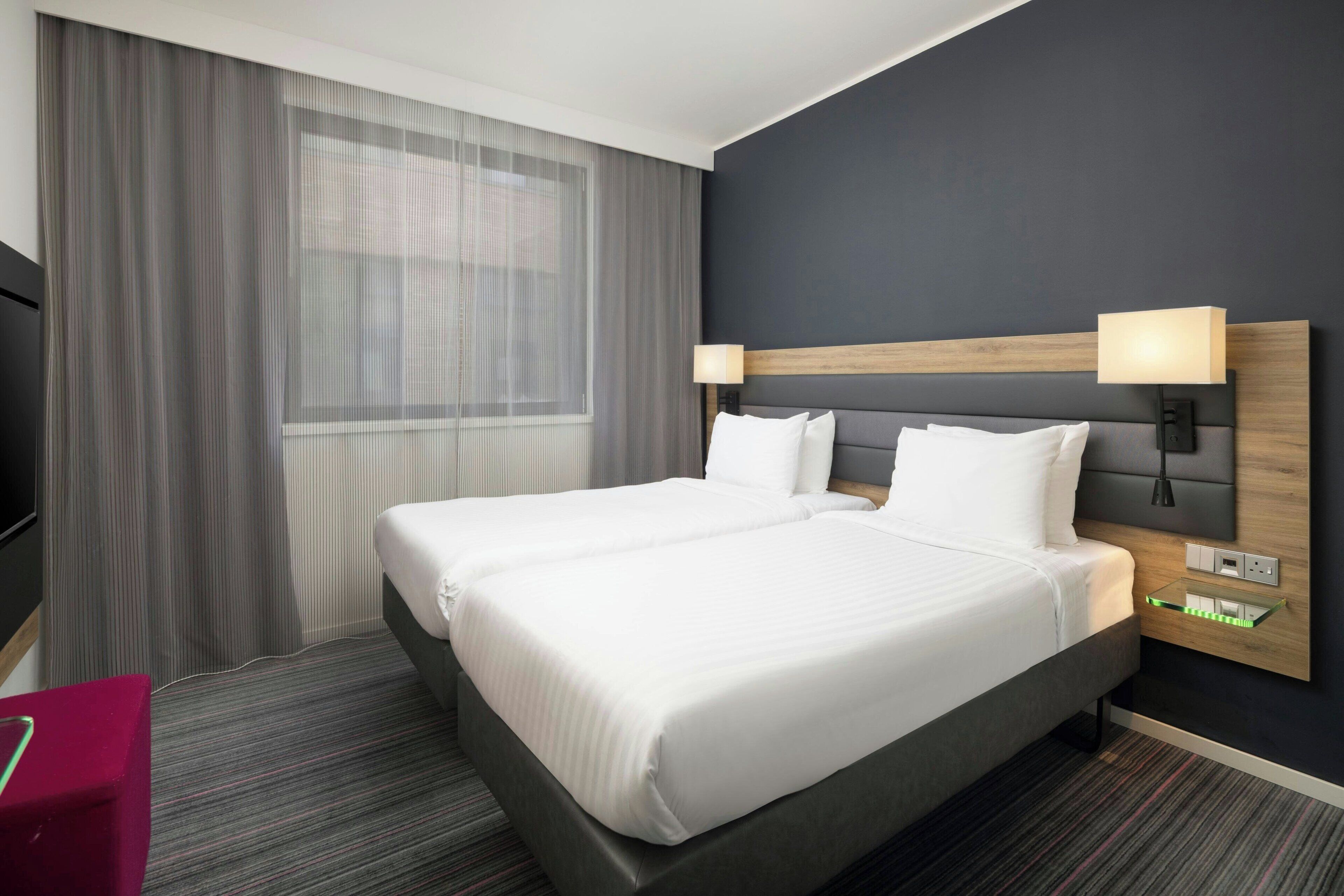 A hotel room with two beds, a TV, a desk with a lamp, and a window with curtains.