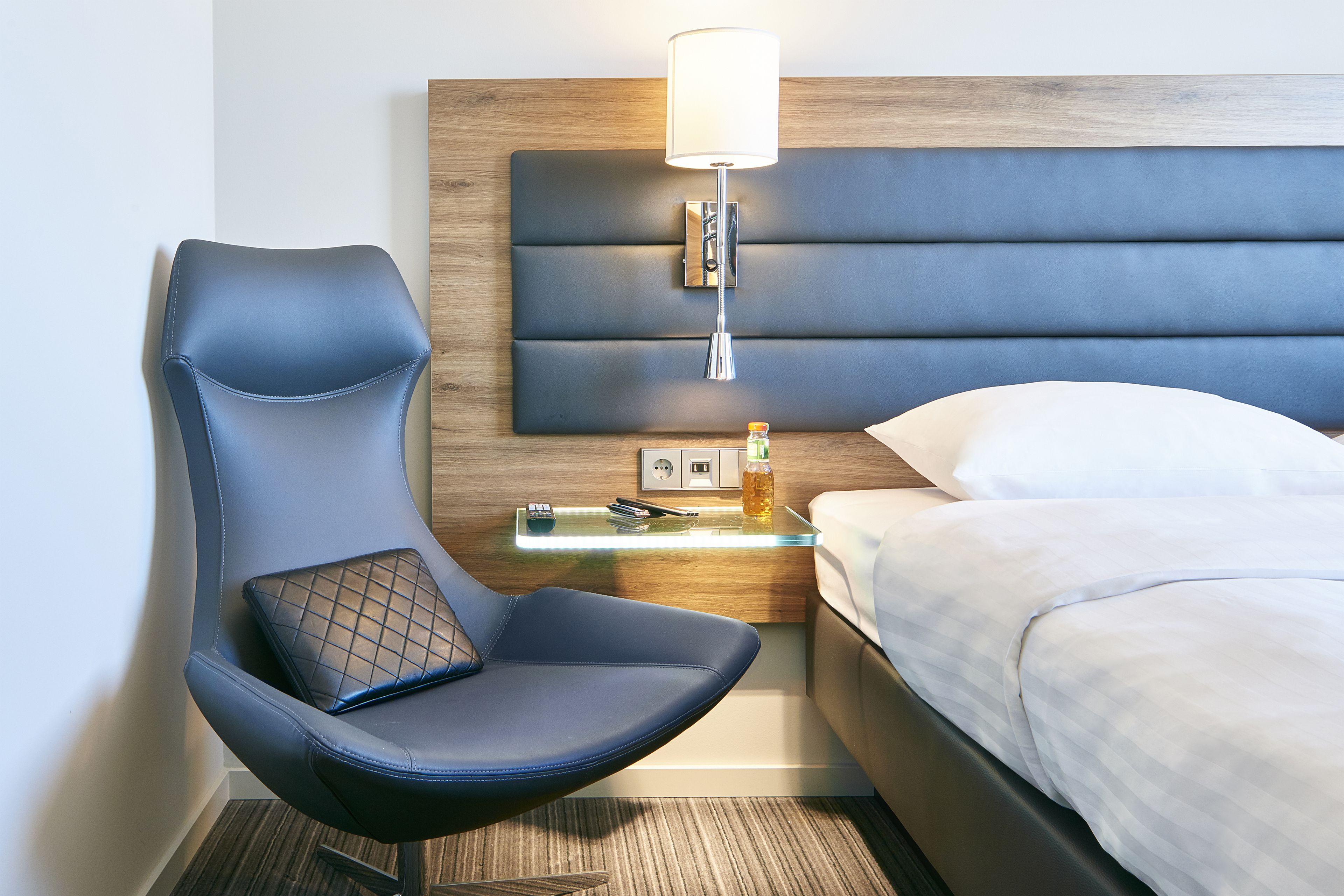 Une chambre d’hôtel moderne avec un fauteuil bleu à revêtement en tissu, une table de chevet avec une lampe et un lit parfaitement fait.