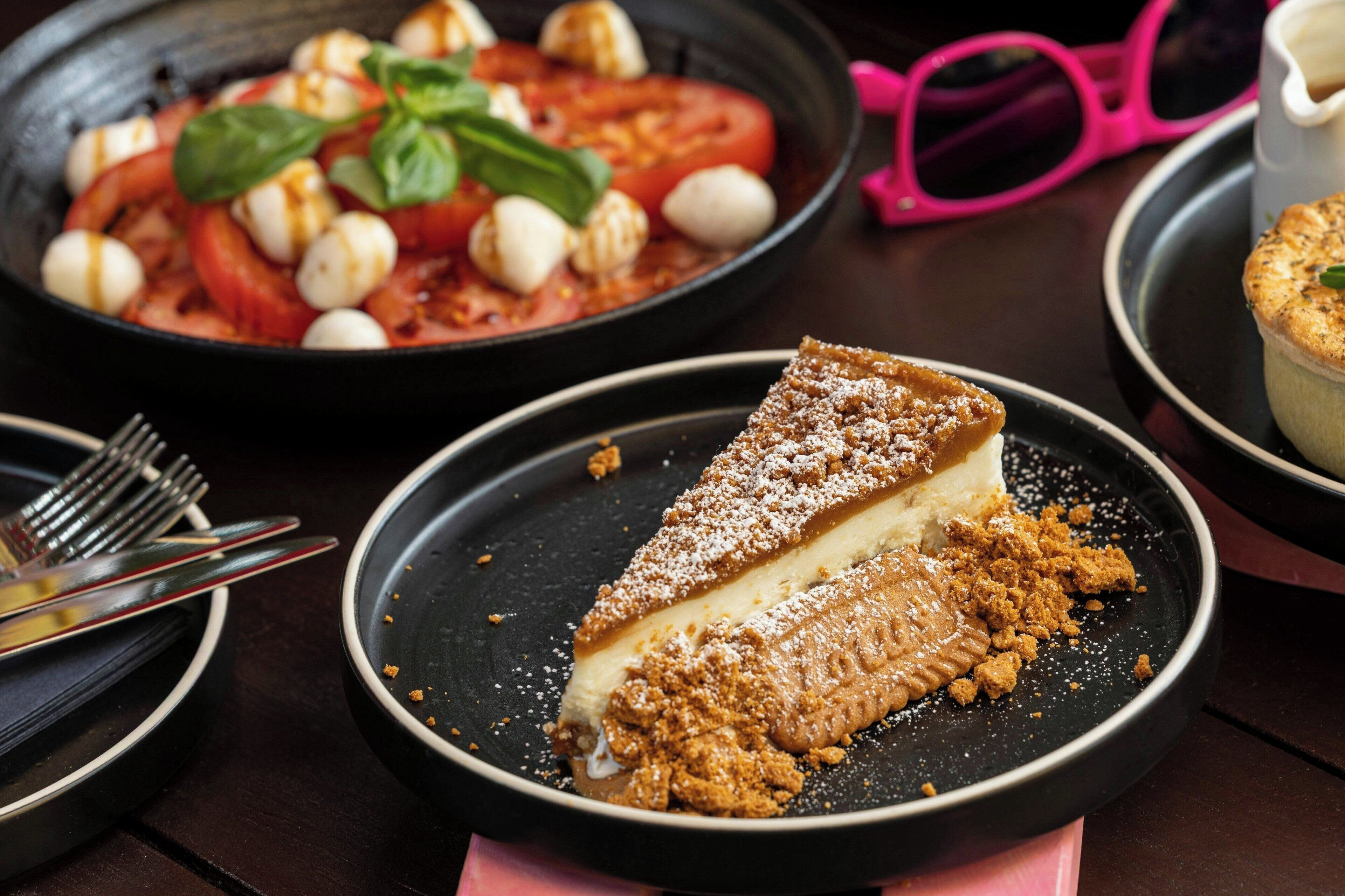 A dessert plate with a slice of cheesecake and powdered sugar, a bowl of tomato salad with mozzarella balls, and a dessert with a crumble topping.