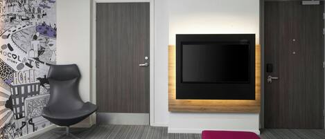 A modern hotel room with a large flat-screen TV mounted on the wall, a black chair, a red ottoman, and a patterned wall mural.