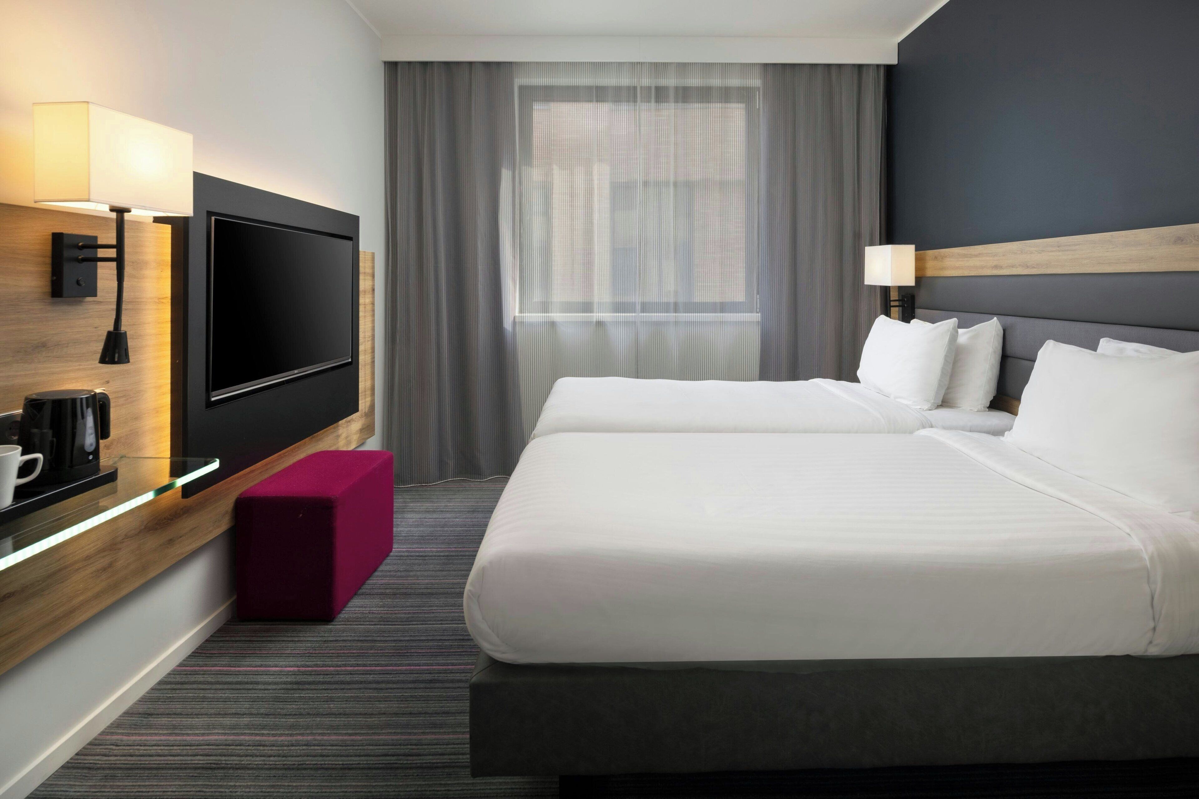 Habitación de hotel con televisión de pantalla plana, pared de madera decorativa, ventana grande con cortinas, cama con sábanas blancas y un pequeño taburete rojo.