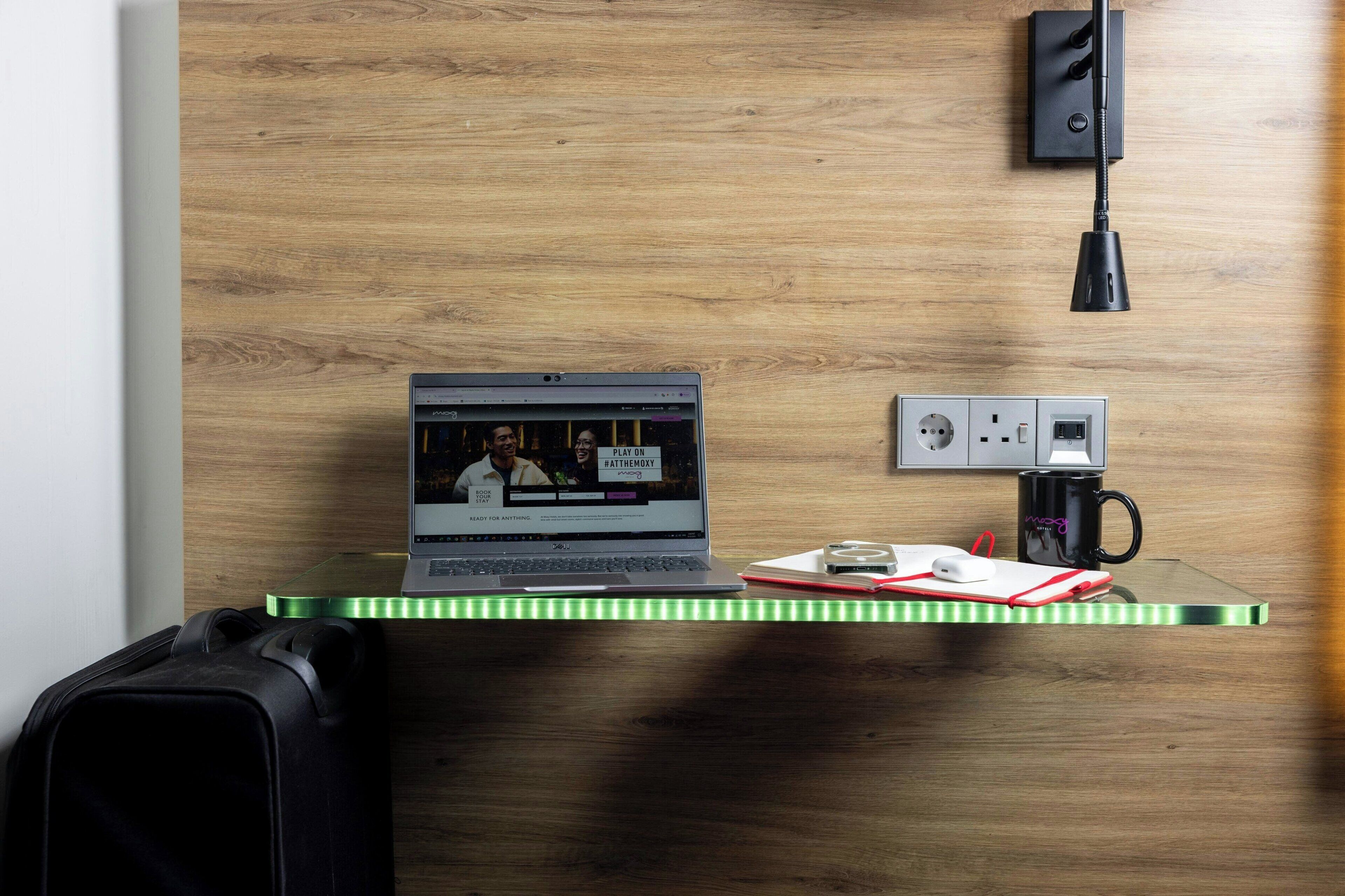 A laptop on a desk with a wooden wall, a power outlet, and a black lamp.