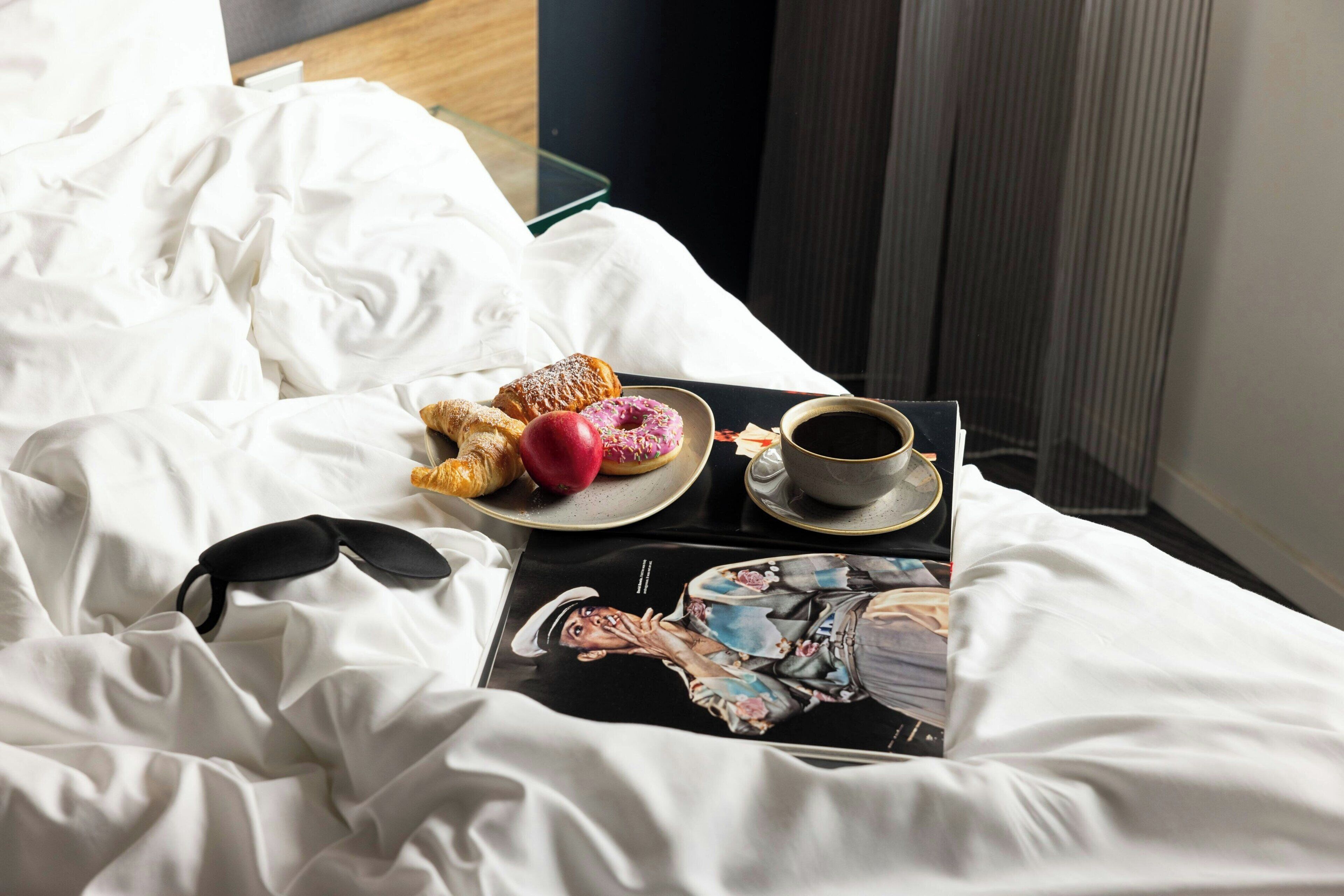 Una cama bien hecha con sábanas blancas, una bandeja con pasteles y una taza de café, y una revista sobre la cama.