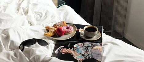 A neatly made bed with white linens, a tray with pastries and a cup of coffee, and a magazine on the bed.