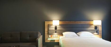 Quarto de hotel com cama, sofá, mesa de centro com cafeteira, controle remoto e mesa de cabeceira com abajur.
