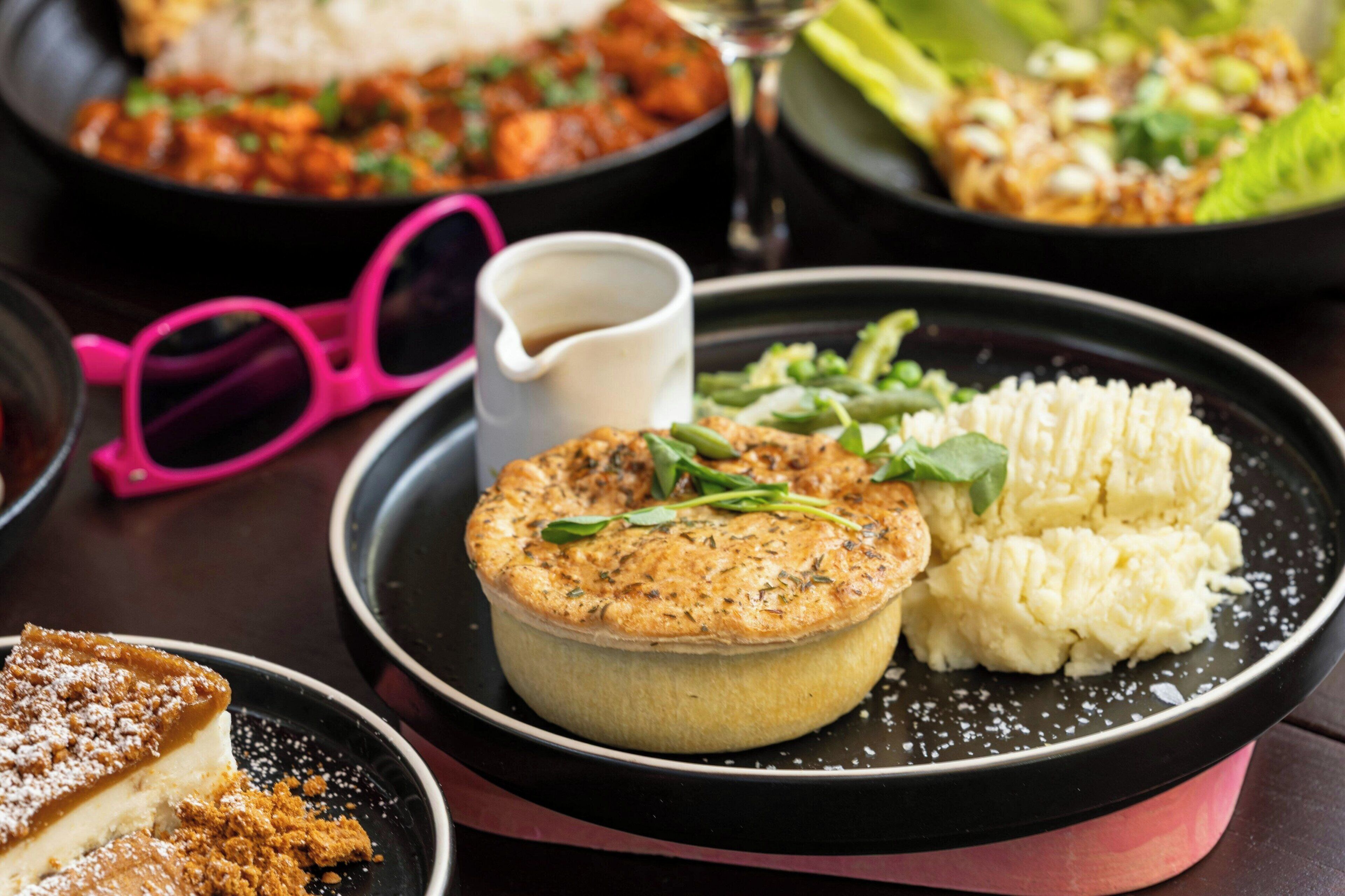 A dessert plate with a pie, mashed potatoes, and greens, accompanied by pink glasses and a small cup.