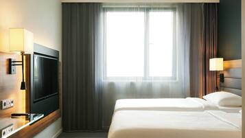Una habitación de hotel con televisión montada en la pared, un escritorio con lámpara, dos camas y un gran ventanal con cortinas.