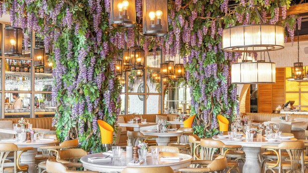 Een restaurant met ronde tafels, paarse bloemen die aan het plafond hangen, en warme verlichting.