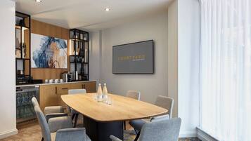 A modern hotel room with a wooden dining table, grey upholstered chairs, a kitchen area with a coffee machine, and a large window with sheer curtains.