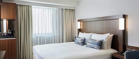 Habitación de hotel con una cama grande, cabecera de madera, escritorio con silla y un ventanal con cortinas.