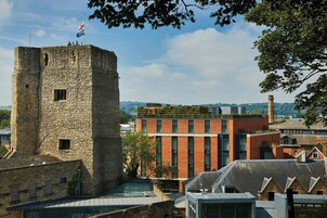 A historic stone tower with a flag, adjacent modern buildings, and a view of the surrounding landscape.