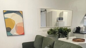 A green sofa with multiple cushions, a modern abstract painting, a potted plant, and a kitchen area with a microwave. - Jacaranda Place - Superior Location  (Plympton)