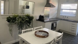 A modern kitchen with white cabinets, a black countertop, a stainless steel range hood, a microwave, and a built-in oven. There is a window with blinds, a hanging plant, and a wooden tray on the table. - Jacaranda Place - Superior Location  (Plympton)