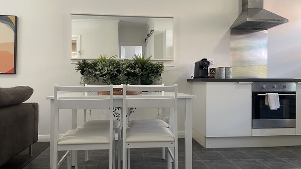 A modern kitchen with white cabinetry, a built-in oven, and a stainless steel range hood. There is a dining table with white chairs and a potted plant on it. - Jacaranda Place - Superior Location  (Plympton)