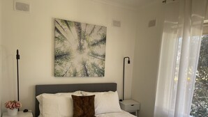 A bedroom with a bed, white bedding, a brown pillow, a nightstand with a vase of flowers, and a painting on the wall. - Jacaranda Place - Superior Location  (Plympton)