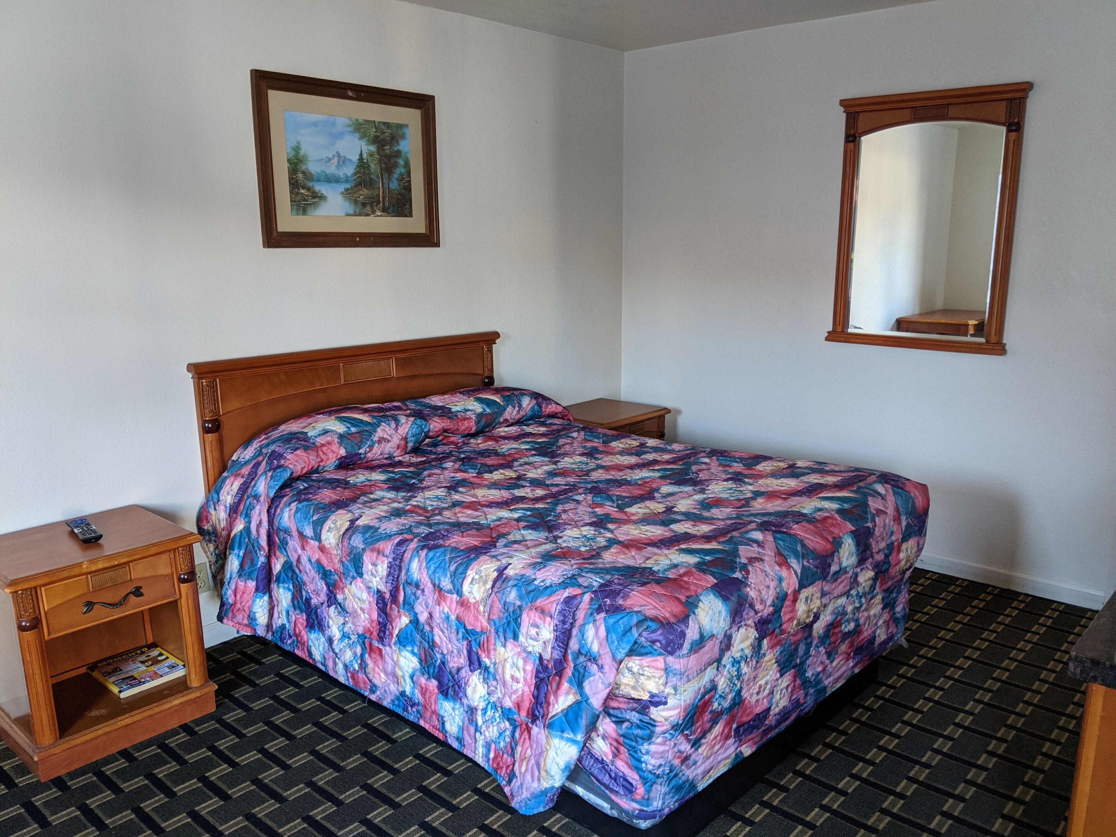 A hotel room with a wooden bed, a nightstand with a remote control, a small table, a framed picture on the wall, and a mirror.