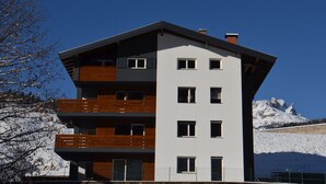 Ein mehrstöckiges Gebäude mit Balkonen, einem geneigten Dach und einer gebirgigen Kulisse.