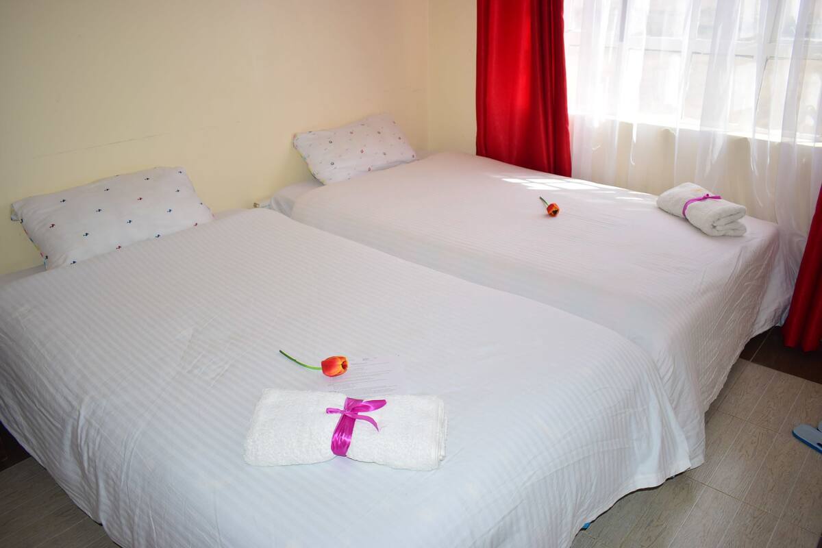 Two single beds with white linens, a red curtain, and a window with sheer curtains.