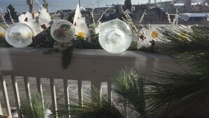 A decorative arrangement of glass spheres and pine branches on a balcony railing overlooking a cityscape with a body of water and a bridge in the distance.