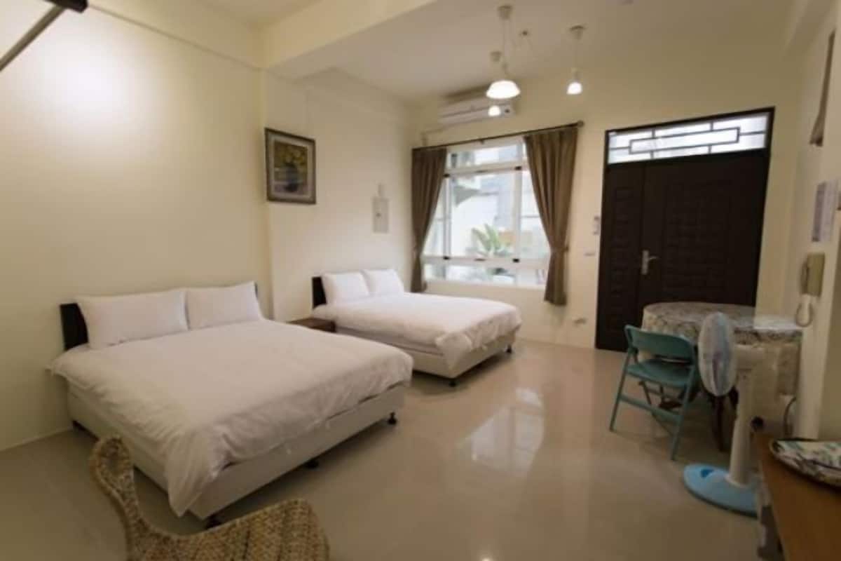 A hotel room with two beds, a small table, a chair, and a window with curtains.