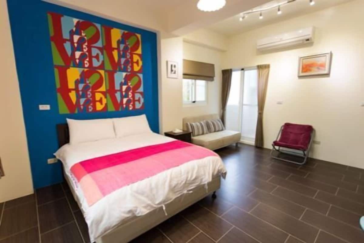 A hotel room with a bed, a chair, a small table, a wall art piece, and a window with curtains.