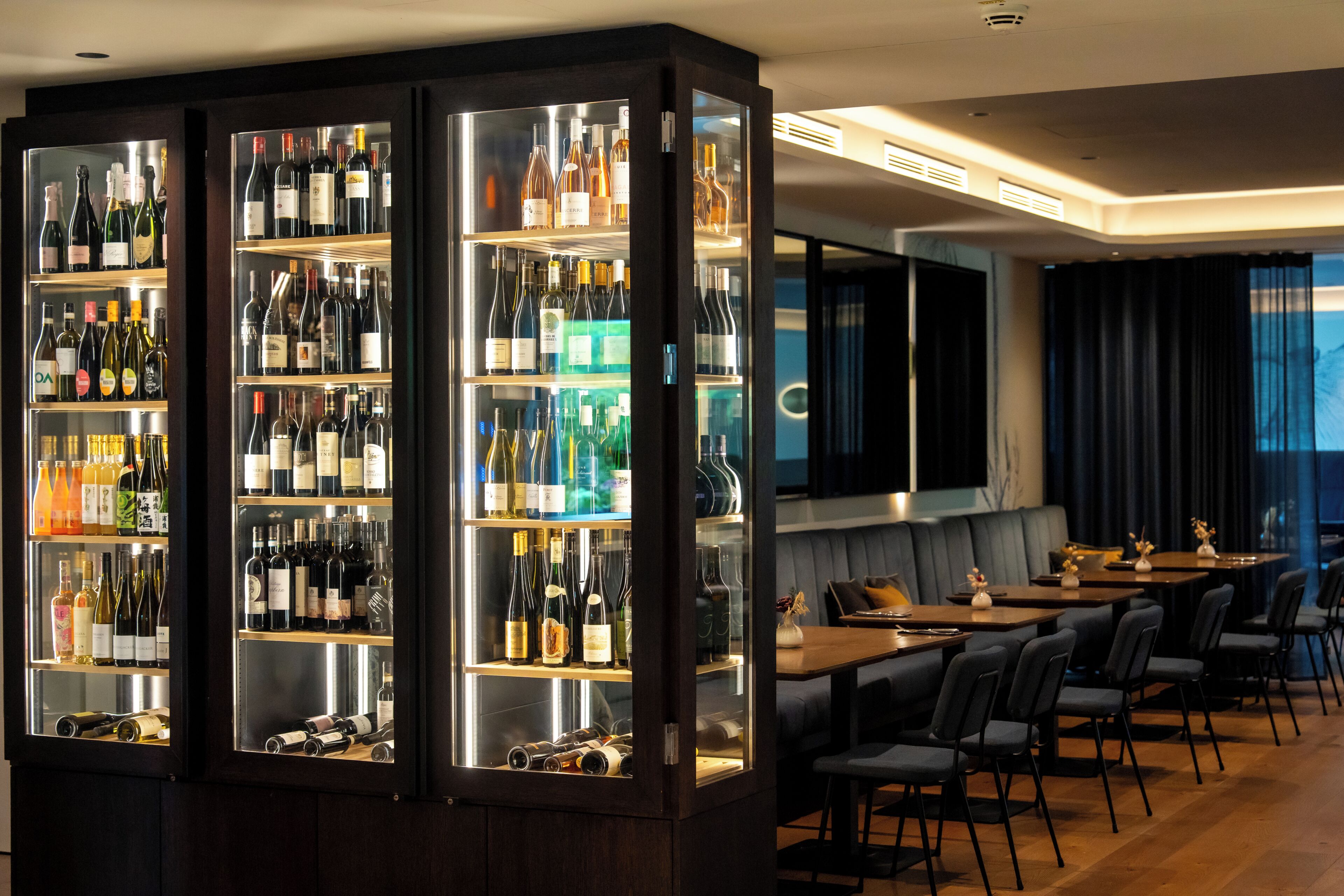 A bar area with multiple shelves of bottles, a seating area with tables and chairs, and a large window with curtains.