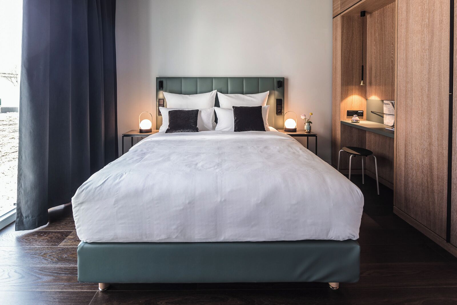 A neatly made bed with a green headboard, two bedside tables with lamps, a small desk with a chair, and a view of the outdoors through a window with dark curtains.