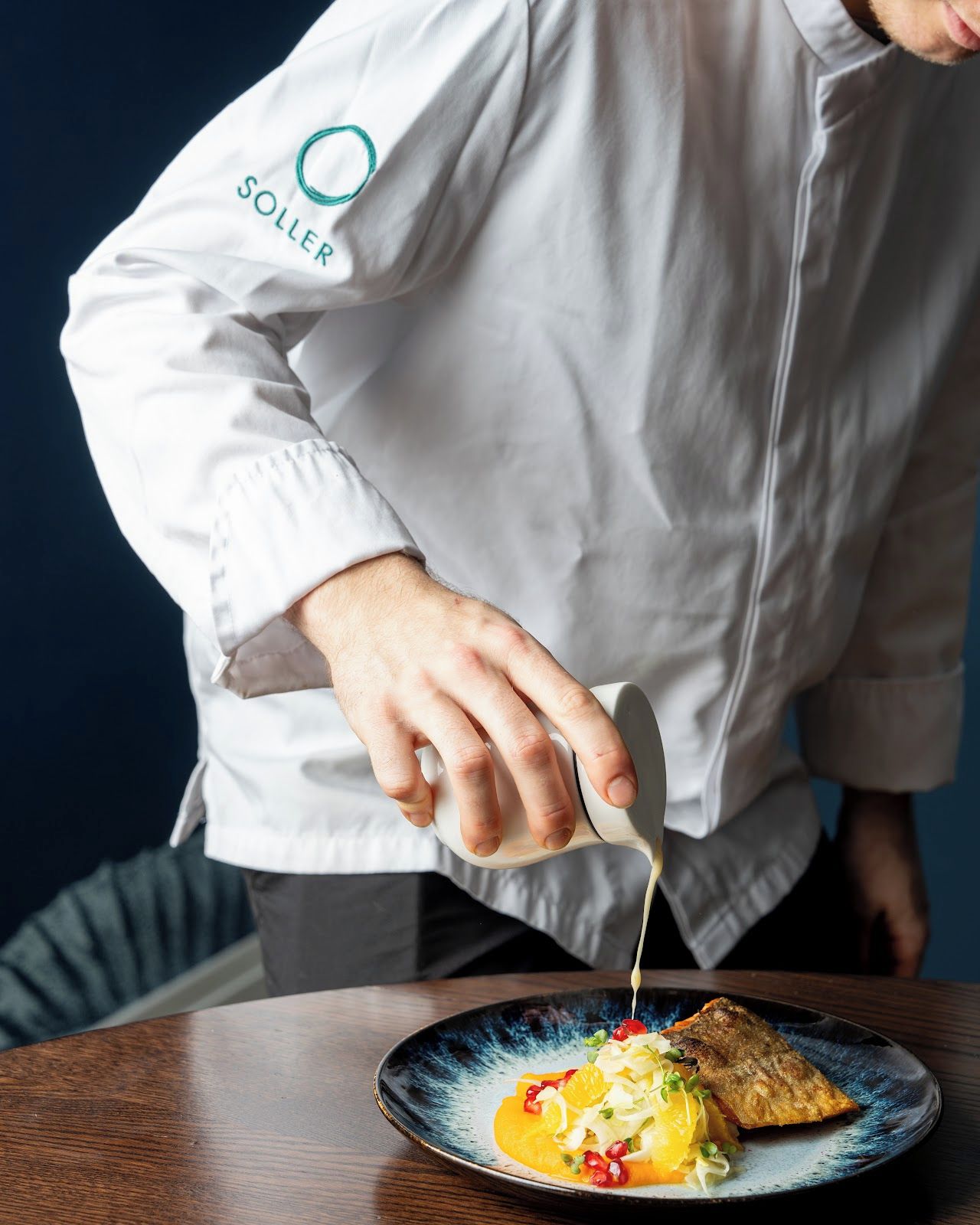 Una persona con delantal de chef vierte salsa sobre un plato de comida.