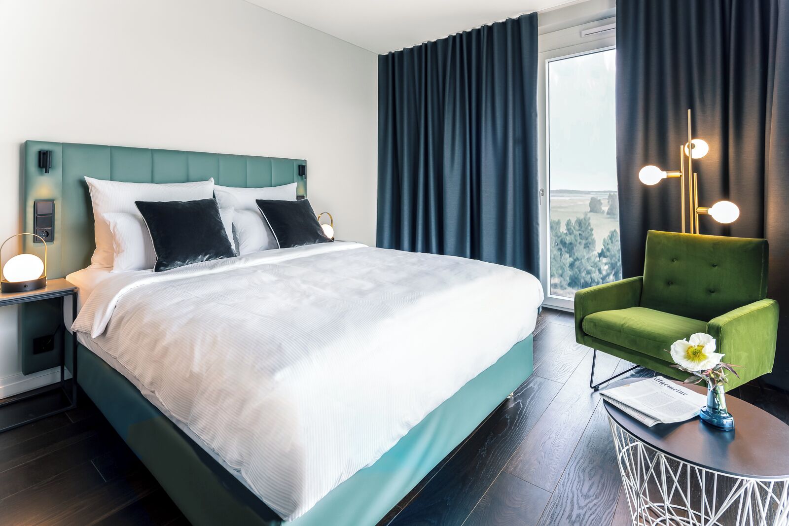 A modern hotel room with a large bed, a green armchair, and a view of the outdoors.