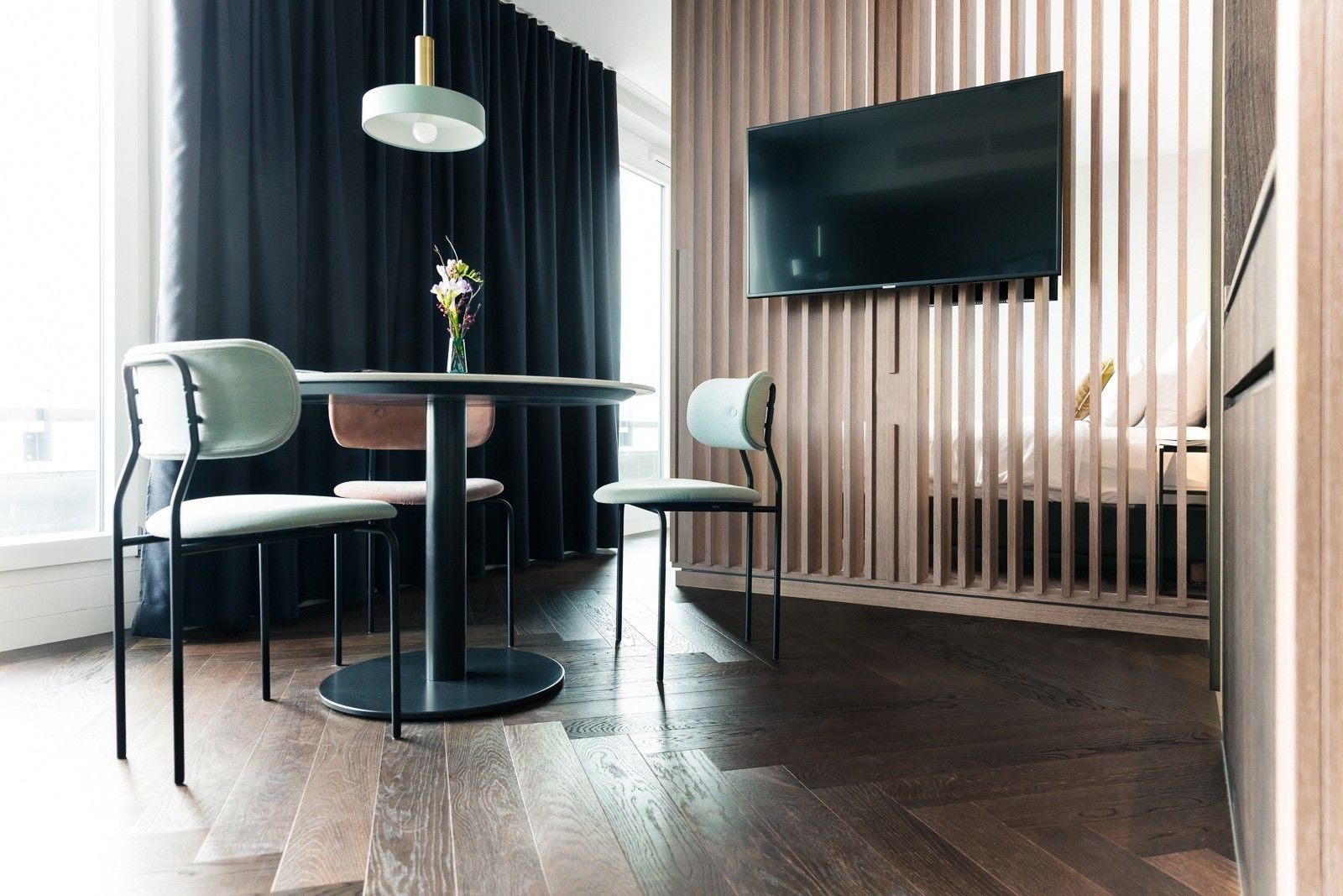 Una sala de estar moderna con una mesa redonda, dos sillas, un televisor de pantalla plana montado en una pared de paneles de madera y una ventana con cortinas oscuras.
