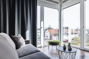 Una sala de estar moderna con un sofá gris, un reposapiés verde y una mesa de centro redonda con jarrones. Amplias ventanas ofrecen vistas a una zona residencial.