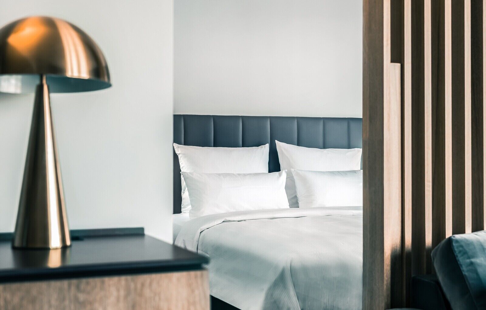 A neatly made bed with white linens, a modern bedside lamp, and a wooden nightstand.