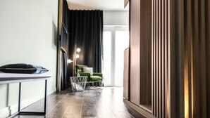 Quarto de hotel moderno com chão de madeira escura, uma poltrona verde, uma pequena mesa branca com um vaso e uma grande janela com cortinas.