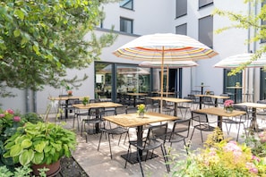An outdoor seating area with tables and chairs, umbrellas, and potted plants. - SOLLER Business Hotel – Munich Airport MUC (Hallbergmoos)