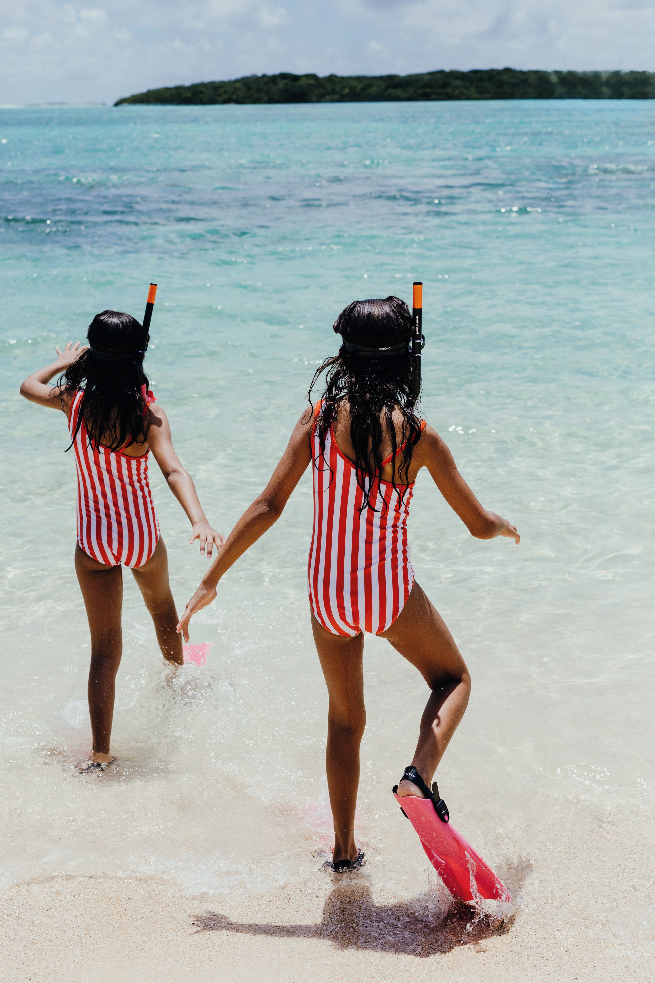 Two individuals in striped swimsuits playing in shallow water with a clear view of the sea and a distant landmass.