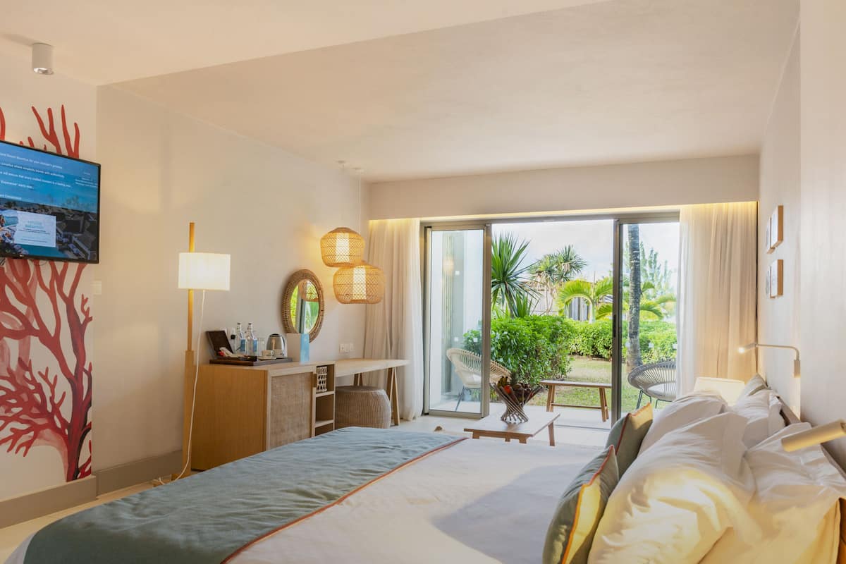 A hotel room with a bed, a desk, a TV, and a view of a garden through the sliding glass door.