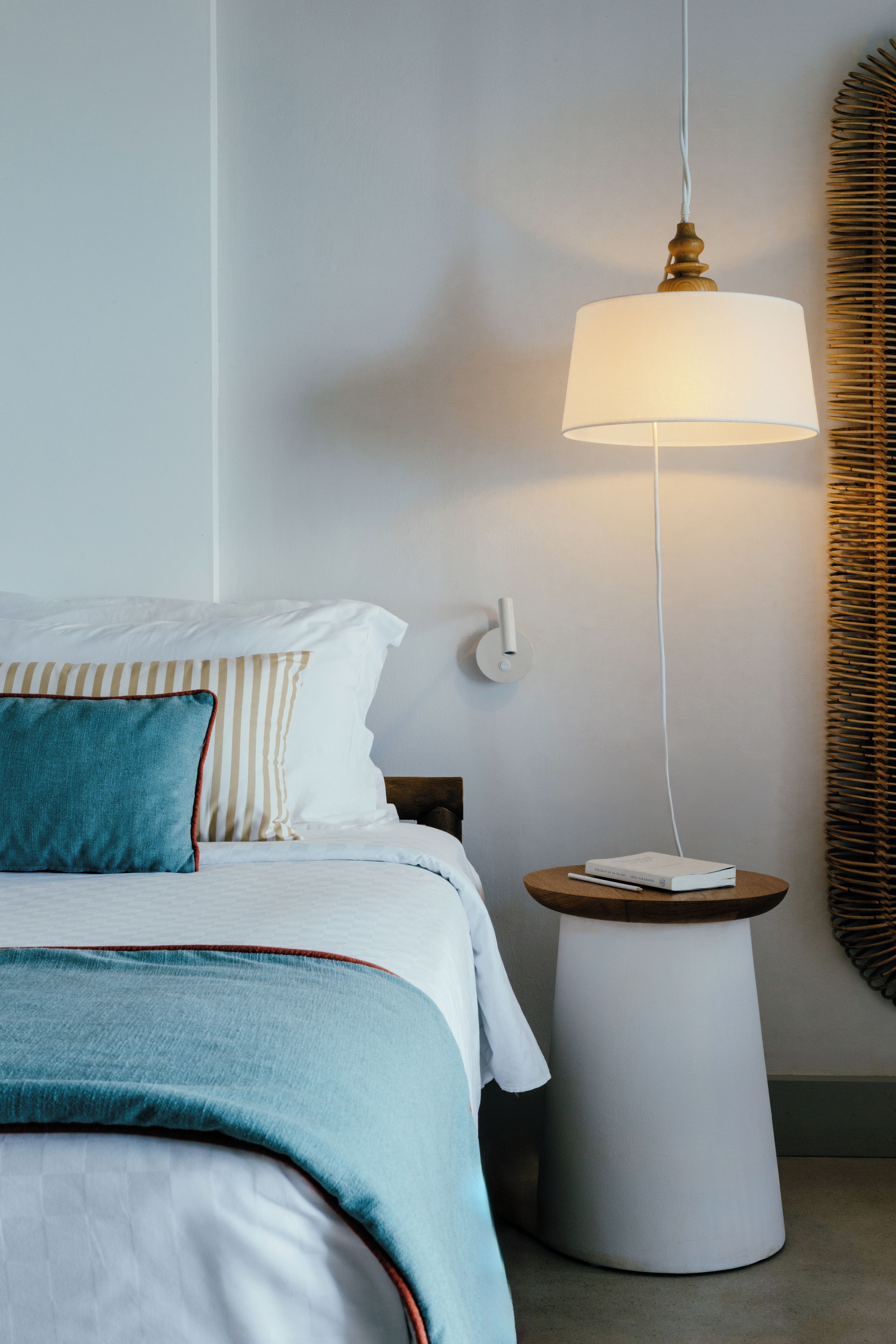 Una cama bien hecha con un almohadón azul y ropa de cama blanca, una mesita de noche con una lámpara y un espejo redondo en la pared.