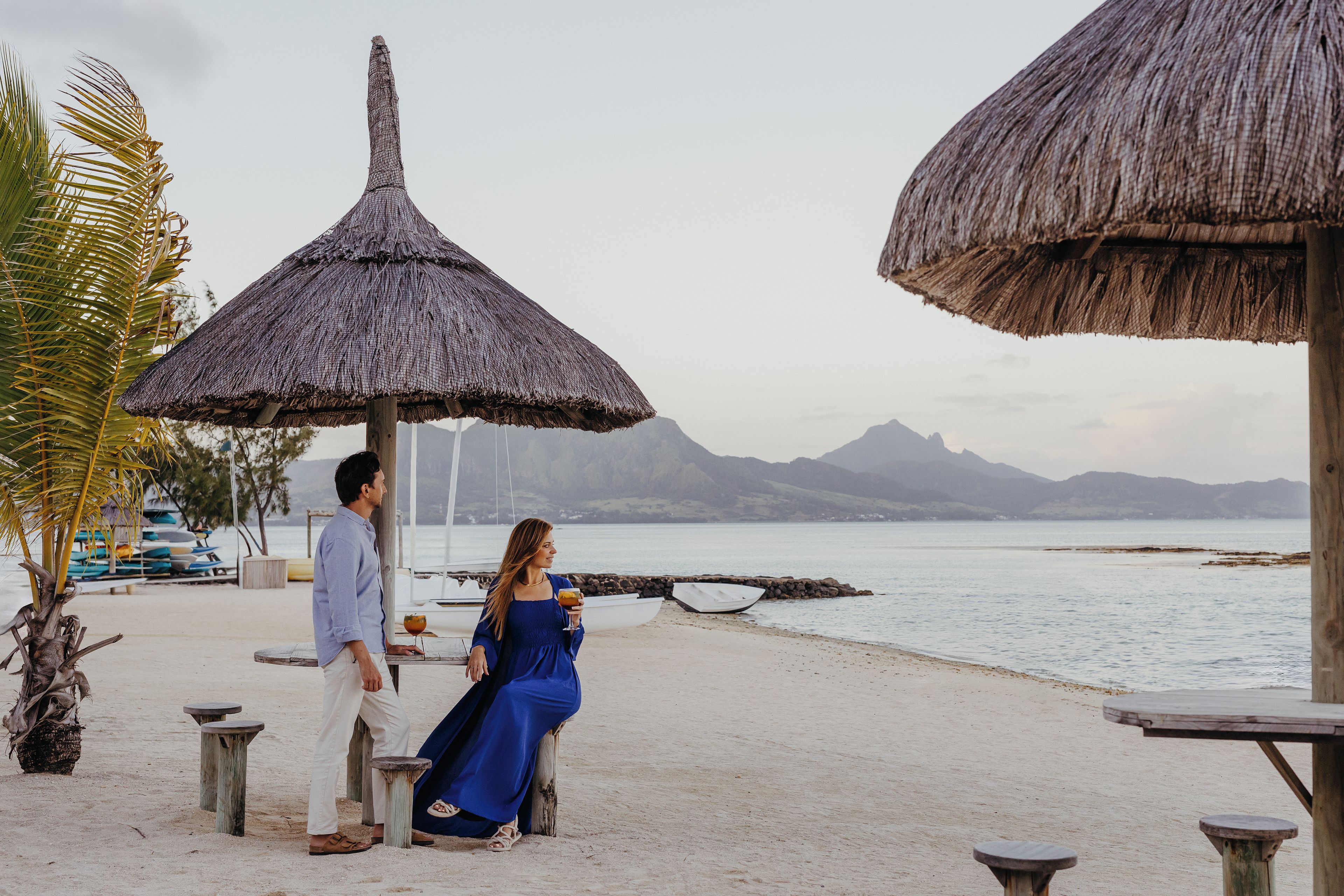 Escena de playa con sombrillas de paja, una pareja sentada y vistas al mar y las montañas.