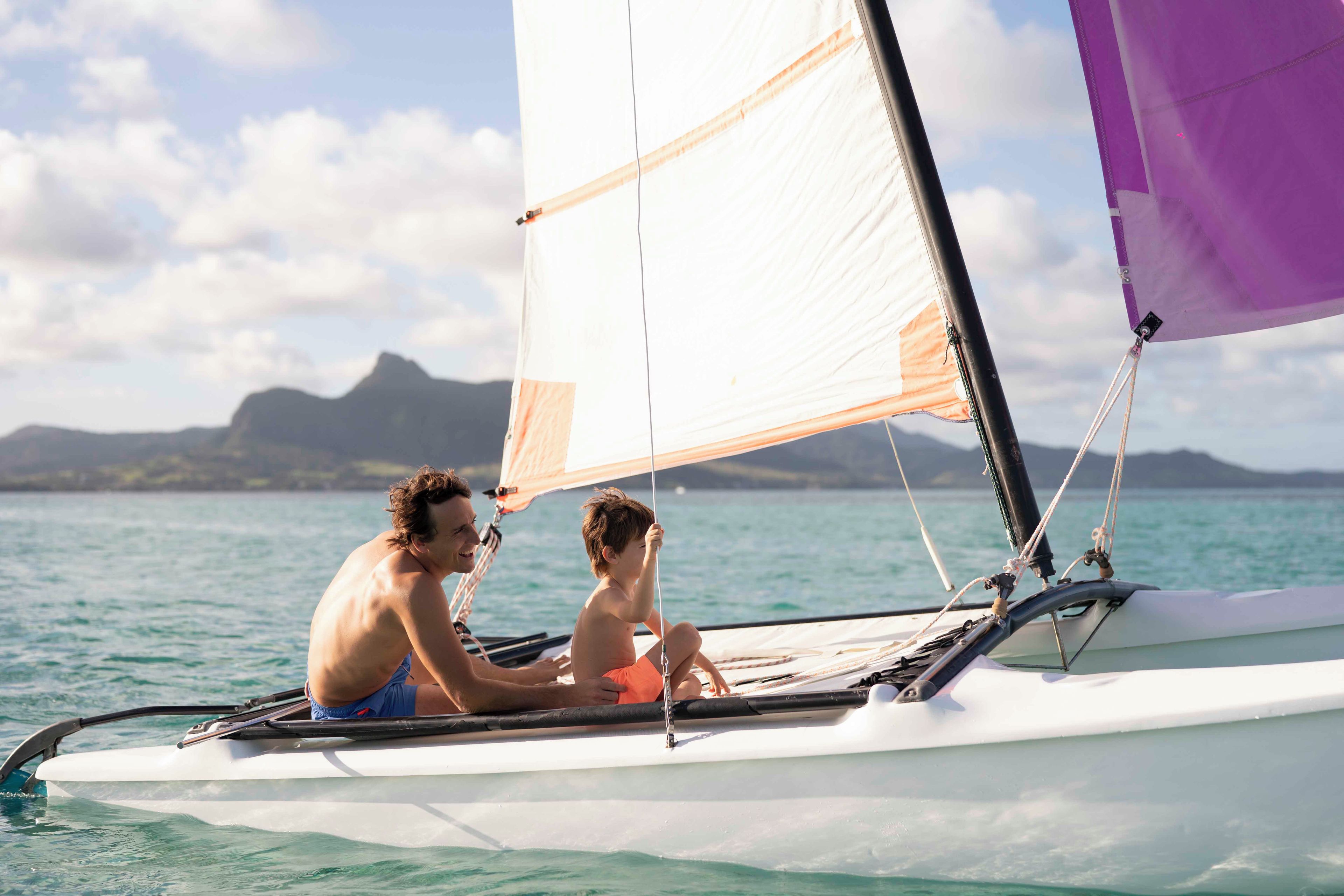 A sailboat with a person and a child on board, sailing on calm waters with a mountainous landscape in the background.