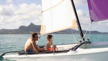 A sailboat with a person and a child on board, sailing on calm waters with a mountainous landscape in the background.