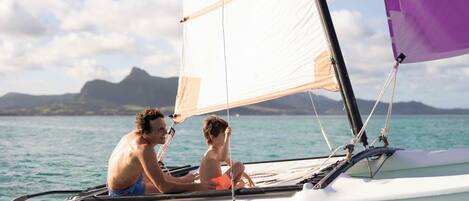 A sailboat with a person and a child on board, sailing on calm waters with a mountainous landscape in the background.