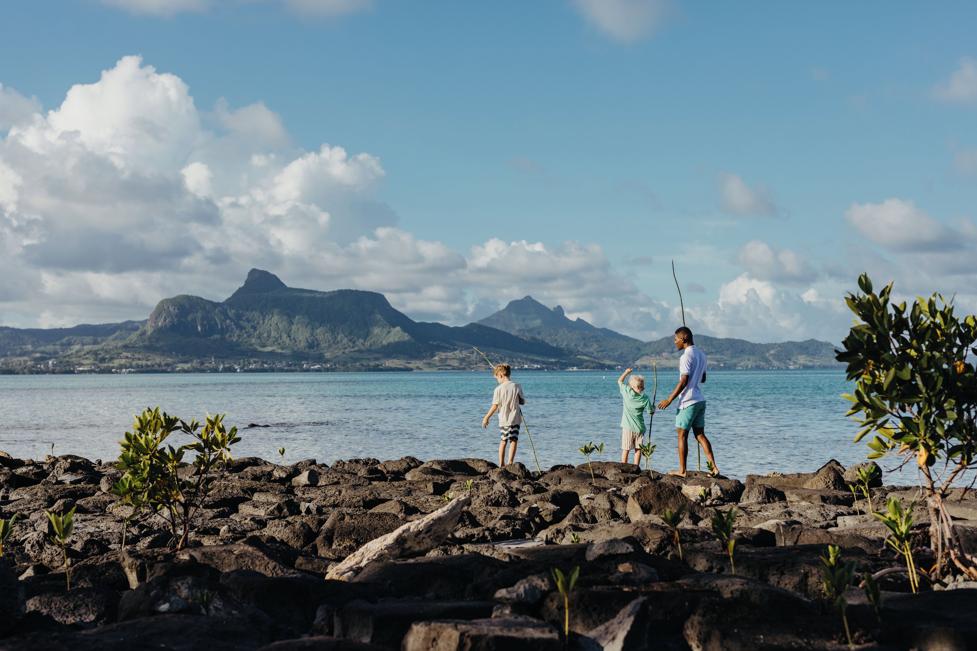 A rocky shoreline with two individuals fishing, a clear sky, and distant mountains.